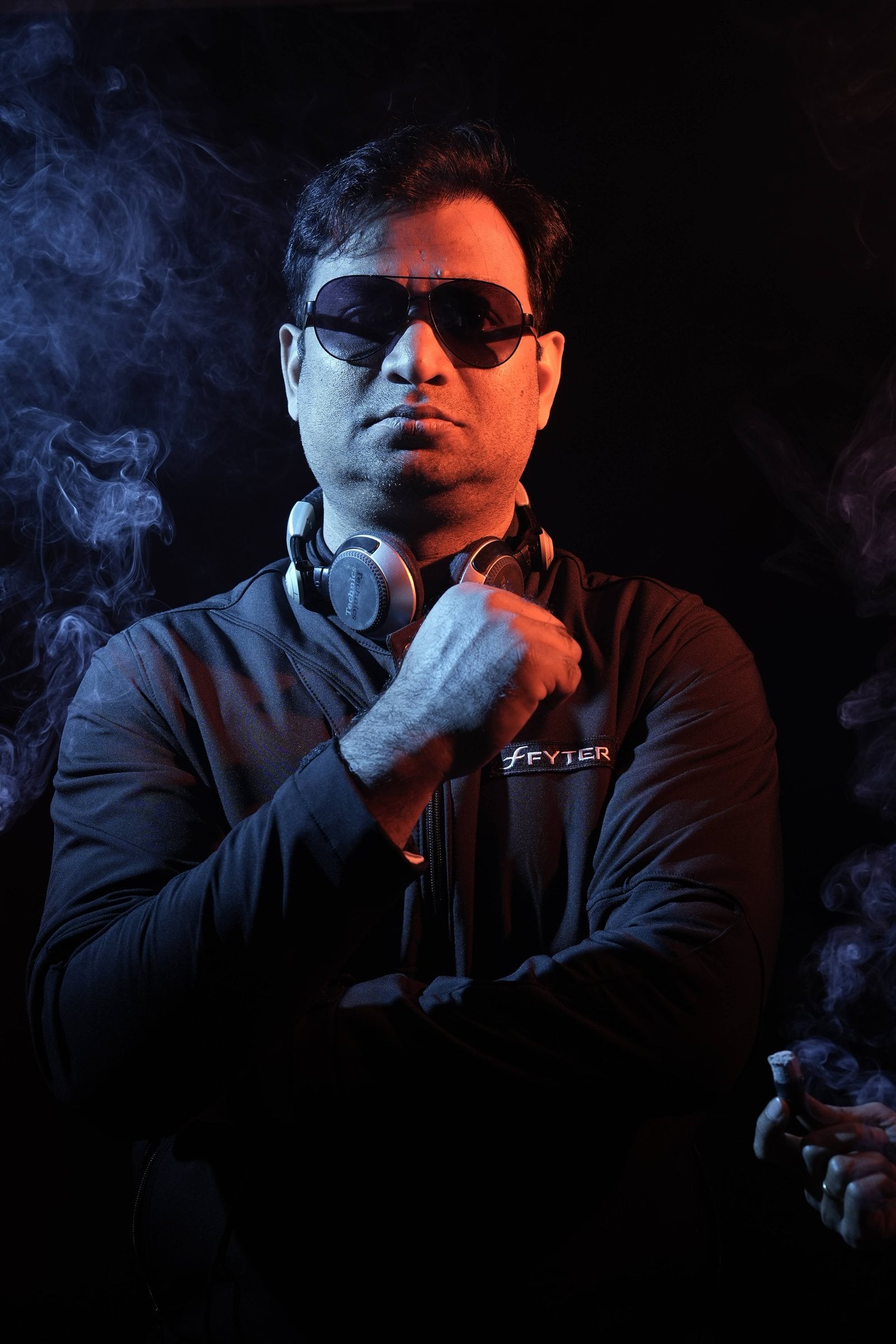 DJ Abdul in dark sunglasses with headphones around his neck, lit in orange and blue through stage smoke