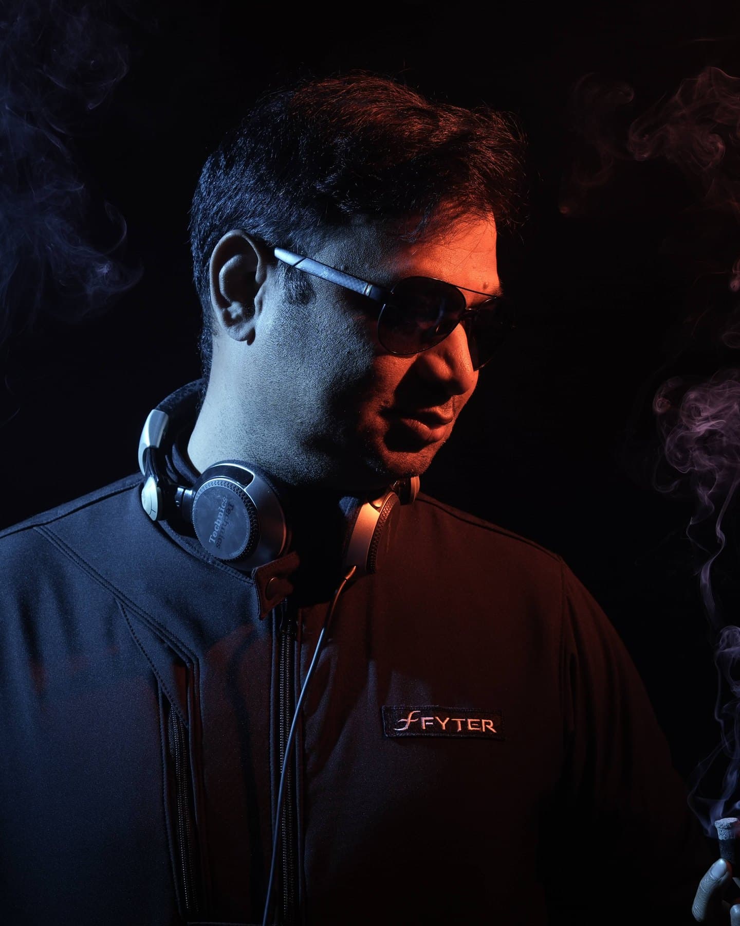 DJ Abdul in sunglasses and headphones, glancing down through orange and blue stage smoke